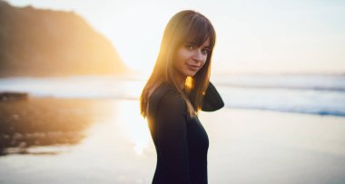 Siyah elbiseli, siyah saçlı, genç ve güzel bir bayan gün batımında kumsalda kameraya bakıyor.