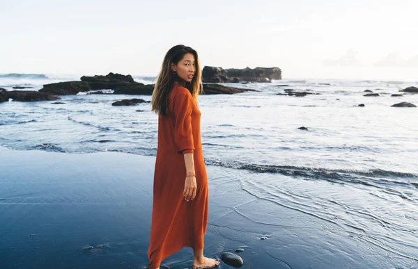 Bali sahilinde kumlu sahil boyunca yürüyen ve yaz tatili boyunca omzunun üzerinden bakan gündelik kıyafetli, düşünceli bir etnik kadın.