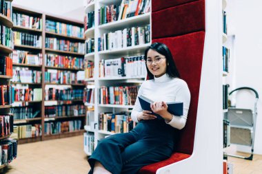 Modern kütüphanede kırmızı döşeme üzerinde otururken gülümseyen ve kameraya bakan oldukça neşeli Asyalı kadın.