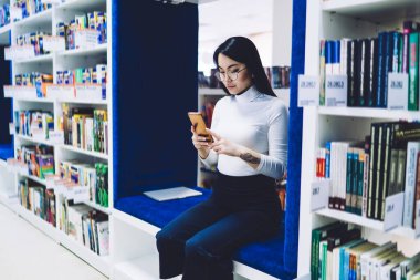 Kitaplıklar arasında mavi kumaşla döşenmiş bankta oturan ve cep telefonundan mesaj okuyan güzel Asyalı genç kız öğrenci.