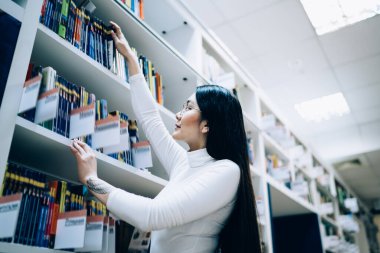 Beyaz kaplumbağa yakalı Asyalı bir kadının kütüphanede dururken üst rafta kitap seçmesinin yan görüntüsü.