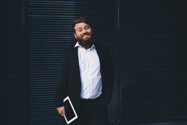 Siyah üniformalı, tablet ve el ele tutuşmuş mutlu sakallı işadamı siyah duvarın yanında duran kameraya bakıyor.