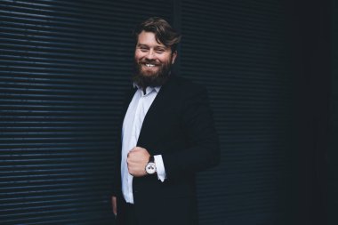 Samimi dişlek, beyaz gömlekli, siyah ceketli, kol saatli, siyah duvarın yanında duran mutlu sakallı adam.