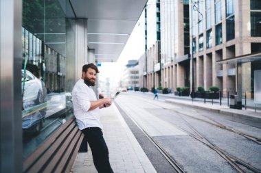 Beyaz gömlekli ve tabletli siyah pantolonlu bir erkek şehir merkezinde tramvay durağında kameraya bakıyor.