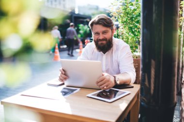 Beyaz gömlekli gülümseyen sakallı girişimci netbook ile çalışıyor tablet cep telefonuyla masada oturuyor ve kağıtlı defter.