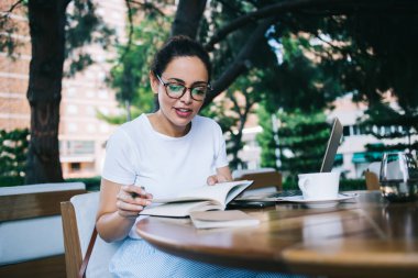 Gündüz kıyafetleri ve gözlükleriyle masada dizüstü bilgisayarı ve çay bardağıyla oturan genç bir kız öğrenci, gün ışığında kitaptan notlar okurken...