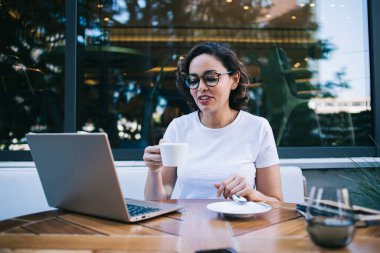 Günlük giysiler ve gözlükler içinde genç bayan çalışan kafenin yanındaki bilgisayarda çalışırken dizüstü bilgisayarı ve bir fincan sıcak içecekle masada oturuyor.
