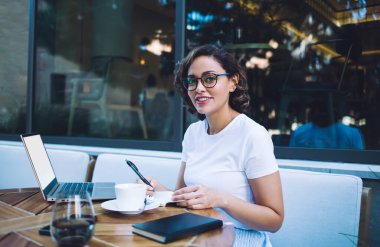 Günlük kıyafetleri ve gözlükleri içinde genç bir kadın bilgisayar ve içki bardağıyla masada oturmuş kafeteryanın yakınındaki kameraya bakıyordu.