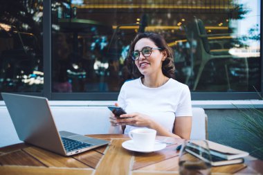 Serbest giyinmiş, gözlüklü, dizüstü bilgisayarı ve sıcak içeceği olan genç bir bayan açık hava kafeteryasında telefonda internette sörf yaparken masada oturuyor.