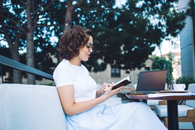 Yan görünüş, şık bir yaz giysisi ve gözlüklü genç bir kadın. Açık kafeteryadaki siyah ekranlı dizüstü bilgisayarla çalışırken günlükte notlar okuyor.