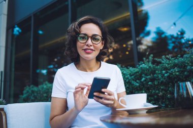 Beyaz tişörtlü, gözlüklü, modern cep telefonlarını karıştıran ve modern açık hava restoranındaki masada otururken kameraya bakan pozitif genç bir kadın.