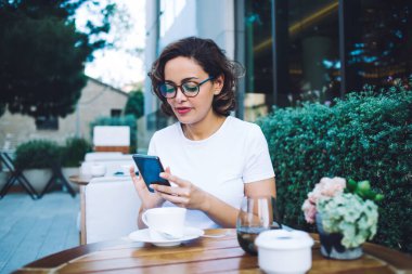 Gözlüklü sakin esmer, çağdaş cep telefonundan mesaj atıyor ve şehir merkezindeki modern kafeteryada yaz gününün tadını çıkarıyor.