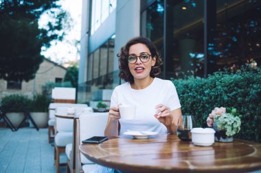 Beyaz gömlekli mutlu bir bayan aromatik taze kahve içiyor ve modern açık hava kafesinde mola verirken kameraya bakıyor.