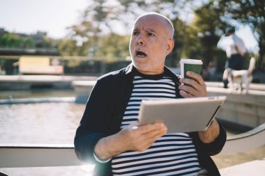 Kahveli ve dijital tabletli hayret verici erkek blogcu güneşli şehirde dinlenen internet haberleriyle şoke oldu. Son sınıf öğrencisinin boş zamanlarında dokunmatik pedi kullanarak yüzündeki ifadeye şaşırdı.