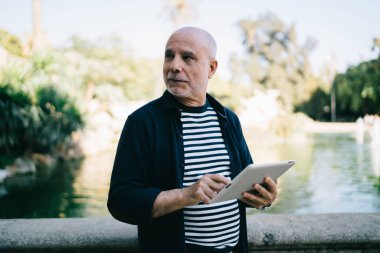 Düşünceli beyaz bir adam elinde dijital tablet tutarken düşünceli bir şekilde kafasını çeviriyor, modern dokunmatik sayfası olan yaşlı bir erkek seyahat metnini düşünüyor.