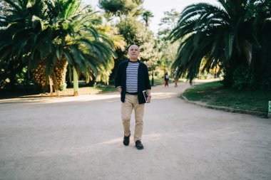 Beyaz bir erkek, emekliliğinde palmiye ağaçlarıyla şehir parkında yürüyor. Düşünmek için boş zamanlarının tadını çıkarıyor. Günlük kıyafetleri içinde modern dokunmatik ped teknolojisini elinde tutuyor ve geziniyor.