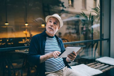 Modern dokunmatik defteri olan düşünceli bir erkek turist kaldırım kenarındaki bankta dinlenirken düşünceli bir şekilde kafasını çevirmiş dijital tabletle kafkasyalı üst düzey bir turist ağ bloglarını düşünüyor.