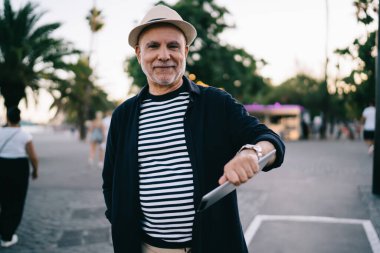 Emekliye ayrılmış modaya uygun giyinmiş erkek turistin yarım boy portresi modern dokunmatik pedi elinde tutuyor ve gündüzleri yürüdüğü için kameraya gülümsüyor, saman şapkalı mutlu beyaz adam sokakta poz veriyor.