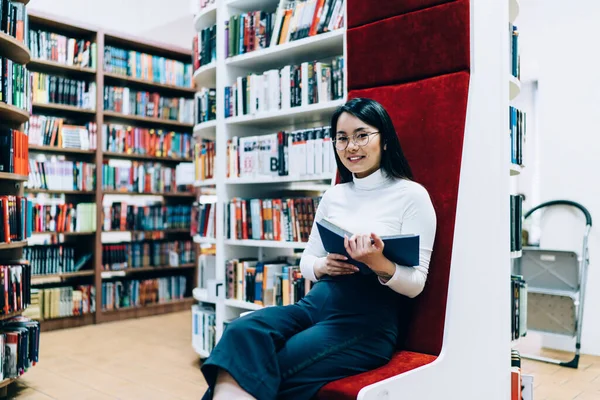 Modern kütüphanede kırmızı döşeme üzerinde otururken gülümseyen ve kameraya bakan oldukça neşeli Asyalı kadın.