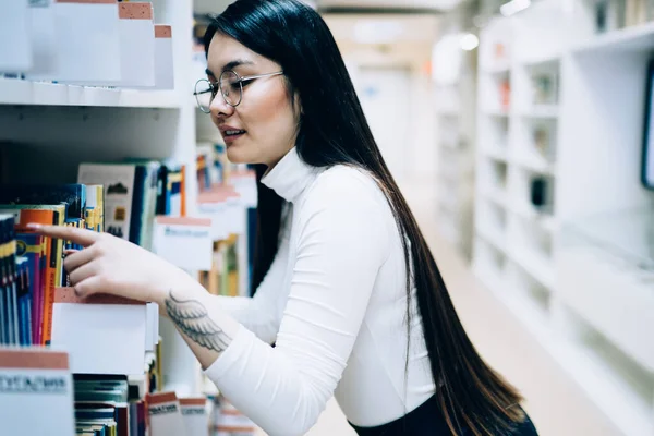 身穿白色上衣 头戴眼镜的黑发年轻亚洲女性在图书馆的书架上拿起了书 — 图库照片