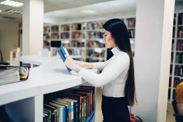 大学図書館の白い湾曲したカウンターに立つ眼鏡に集中したアジア系女子学生の側面図と読書本 — ストック写真