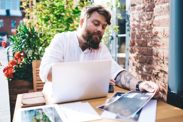 Beyaz gömlekli, konsantre sakallı erkek girişimci açık hava kafesindeki masasında kağıt ve aletlerle çalışıyor güneş ışığında çiçek açan çiçeklerle.