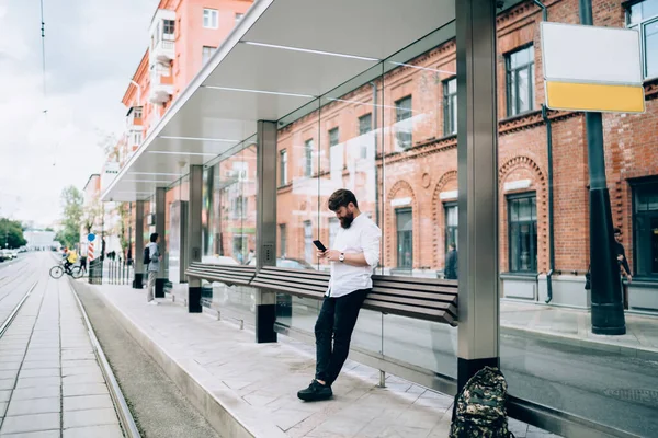 Sırt çantalı yetişkin bir adam tramvay istasyonunda duruyor ve bulutlu bir günde boş şehir caddesinde tuğladan binalara karşı akıllı telefonlara bakıyor.
