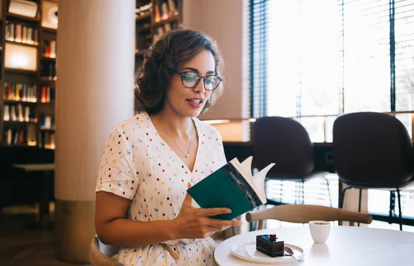 Açık renk elbiseli romantik bir kadın kütüphanede bir parça kek ile oturmuş kitap okuyor ve ara veriyor.
