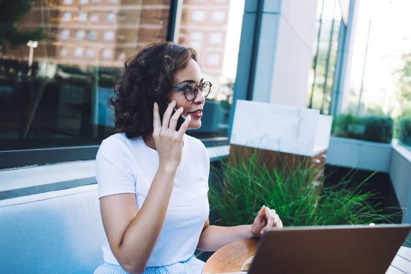 Gündelik giysiler ve gözlükler içinde genç bir kız öğrenci. Masada dizüstü bilgisayarla oturuyor ve açık bir kafede cep telefonuyla konuşuyor.