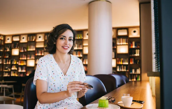 Kitapçıda kameraya bakarken masada oturan tatlı kremalı kek ve bir fincan kahveyle samimi bir bayan.