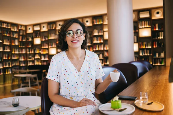 Bir fincan sıcak içecekle ve kitapçının kahve dükkanındaki kameraya bakan lezzetli kremalı pastayla gözlüklü, neşeli genç bir kadın.
