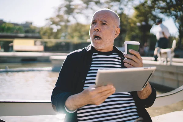 Kahveli ve dijital tabletli hayret verici erkek blogcu güneşli şehirde dinlenen internet haberleriyle şoke oldu. Son sınıf öğrencisinin boş zamanlarında dokunmatik pedi kullanarak yüzündeki ifadeye şaşırdı.