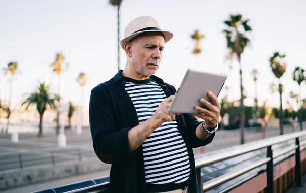 Hasır şapkalı beyaz adam solo seyahat sırasında online rezervasyon yapıyor ve yemek pişiriyor, yaşlı erkek turist emekli olurken sosyal medyayı tarıyor ve modern dokunmatik ekranda 4G kablosuz kullanıyor.