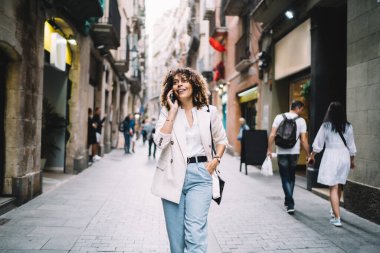 Şık kıyafetler içinde gülümseyen kadın turist şehir caddesinde yürüyor ve uluslararası akıllı telefon konuşmaları sırasında tek başına tatil deneyimini tartışıyor. Gezen mutlu kız hücresini konuşmak için kullanıyor.