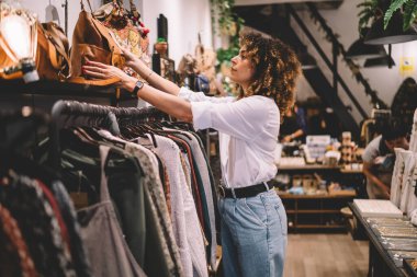 Moda mağazasında kıyafet seçen çekici modacı kadın alışveriş için karar vermeye odaklanmış, yetenekli tasarımcının galerisinde şık kıvırcık kadın kıyafetleri giyip moda modasına uygun bir görünüm yaratıyor.