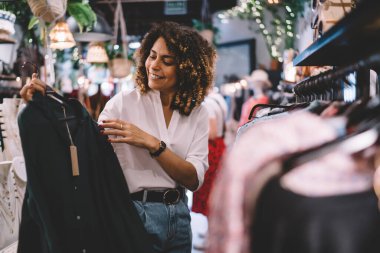 Marka giysileriyle askıların yanında dikilen ve indirimli alışveriş yaparken gülen bir kadın. Komik hippi kız gardırop günlerini butikte geçirdiği günlerin keyfini çıkarıyor.