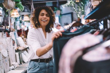 Marka galeride hafta sonu alışverişi yapan neşeli bayan müşterinin portresi, şık kıyafetler giyen neşeli kadın ve askılı elbise giyerken kameraya gülümseyen kadın.