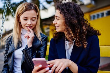 Parkta salıncakta birlikte oturan ve birlikte gülen akıllı telefon ekranına bakan pozitif genç bayan arkadaşlar.