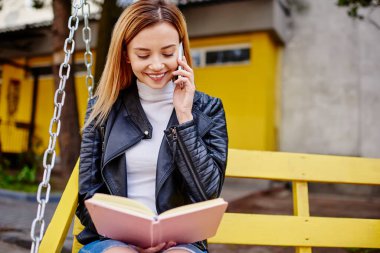 Siyah deri ceketli neşeli bir bayan parkta salıncakta oturuyor ve telefonda konuşurken günlükte notlar okuyor.