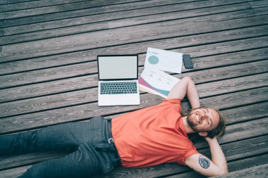 Yukardan gelen neşeli, rahat, günlük giysiler içinde genç bir adam. Parktaki tahta tribünde dizüstü bilgisayar kullandıktan sonra hayal kuruyor.