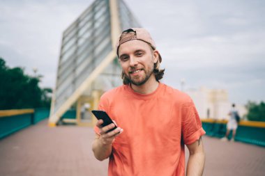 Neşeli, güler yüzlü, sıradan giyinmiş, şapkalı bir erkek sosyal medyada gezinmek için telefon kullanıyor.