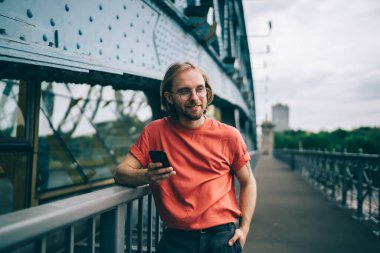 Gülümseyen kızıl sakallı, gözlüklü, uzun saçlı, kırmızı tişörtlü, köprüde cep telefonuyla mesajlaşan ve parmaklıklara dayanan bir adam.