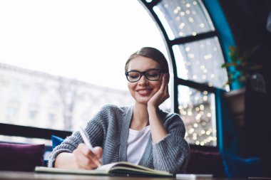 Düşük açılı, dişlek gülüşlü, elleri yanağında masada oturan ve not defterine notlar yazan pozitif kadın.