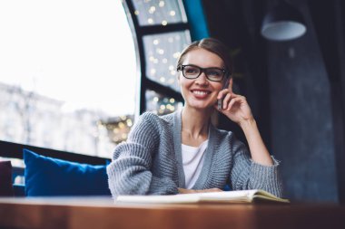 Gündüz vakti rahat bir kafede çalışırken gözlüklü, rahat bir kadının akıllı telefonuyla sohbet etmesi.