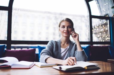 Uzaktan kumandalı genç bir kadın, açık not defteriyle masada otururken ve akıllı telefondan konuşurken gözlerini kaçırıyor.