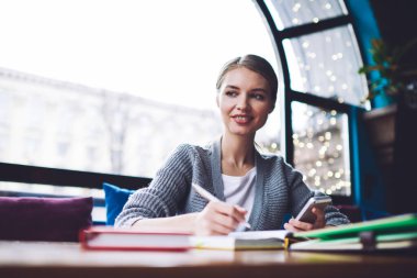 Pozitif kadın serbest çalışanın elinde not defterleri ve akıllı telefonuyla hafif çalışma alanında uzaktan kumandayla otururken başka tarafa bakmasından.