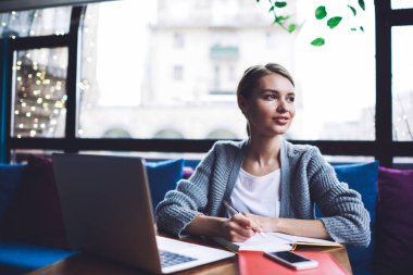 Gündüz vakti geniş pencereli modern çalışma alanında çalışırken, günlük giysiler içinde, dizüstü bilgisayarla tahta masada oturan ve not defterine notlar yazan sakin kadın girişimci.
