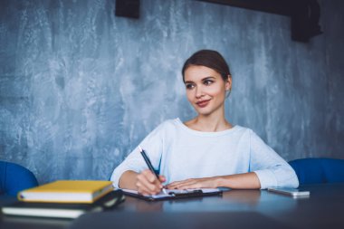 Gündelik giysiler içinde, elinde defterlerle masada oturan ve mavi duvarlı modern çalışma alanında çalışırken dokümanlara yazı yazan pozitif bir kadın işçi.
