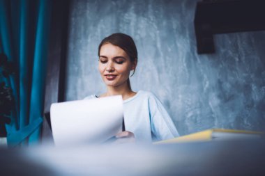 Modern çalışma ortamında masada otururken kahverengi saçlı, belgelere bakan düşük açılı genç bayan öğrenci.