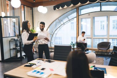 Çağdaş çalışma ortamında parlak lambalar altında birlikte çalışırken, tanınmayan bir partnerle masa başında projeyi tartışan bir grup neşeli çoklu etnik iş arkadaşı.